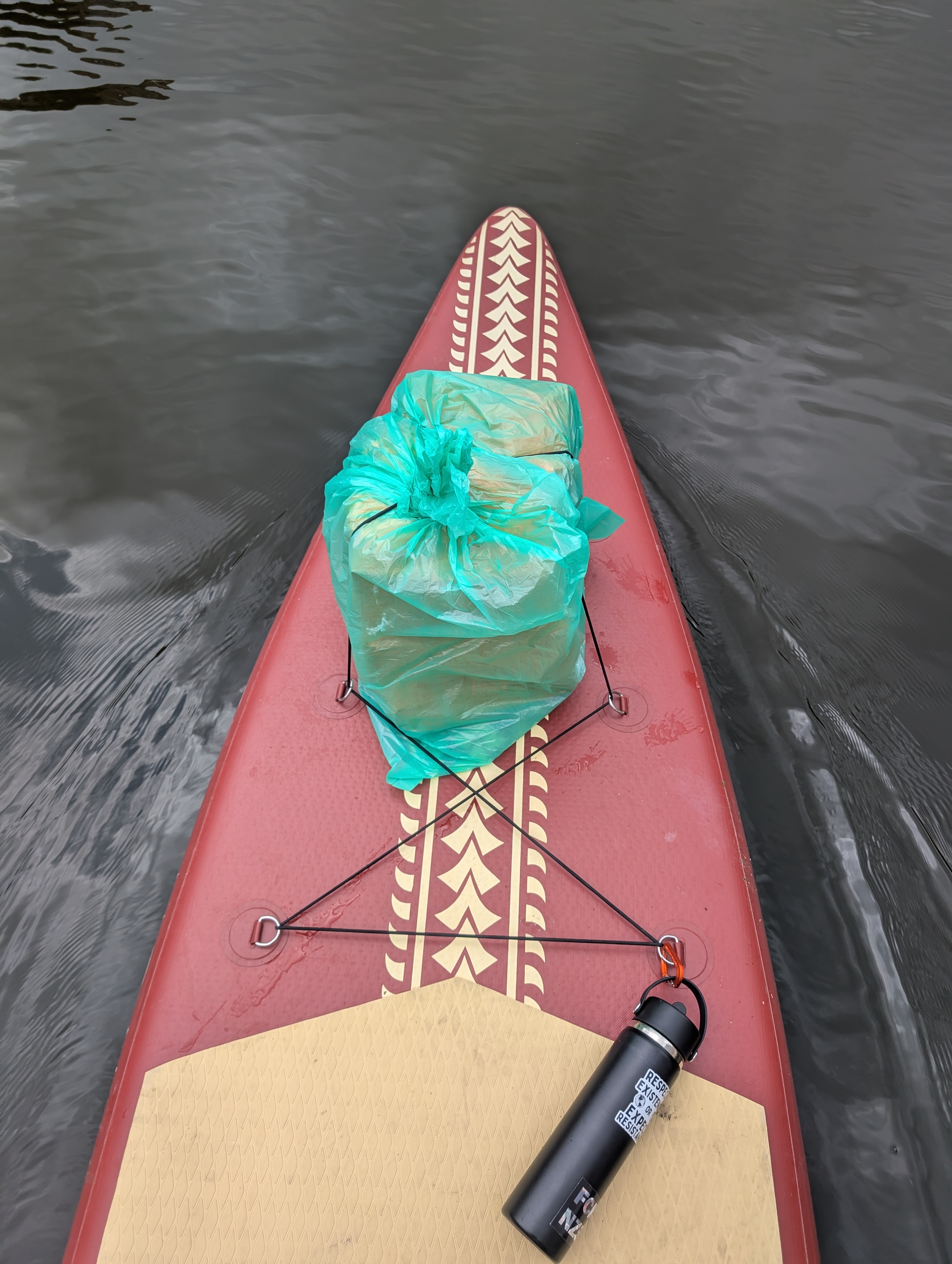 the front of a reddish brown SUP with tan polynesian-style markings in the water. there are ripples around the sup, and some water drops on the surface of the sup. there is a green bag of brown packages tied down on the front