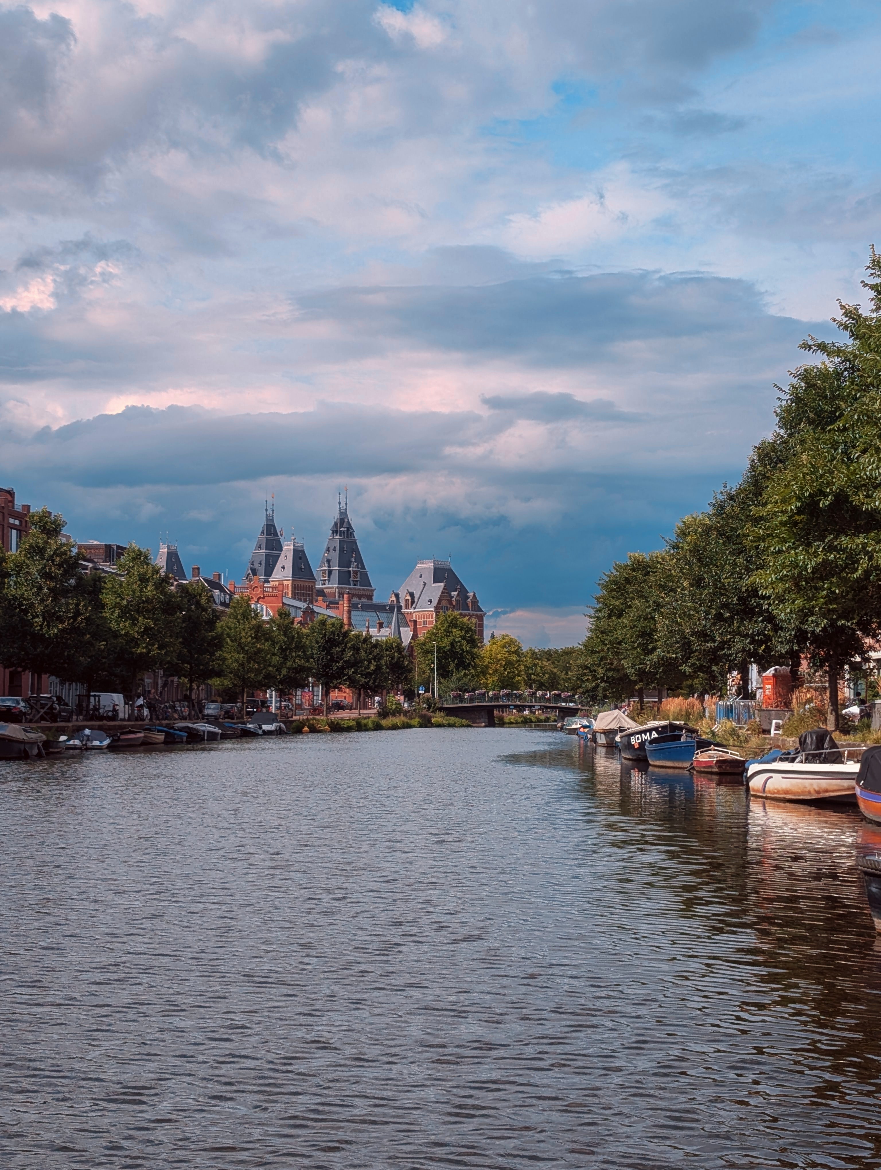 Monday, July 21 2025: The bottom third of the image is canal water with ripples reflecting the sky color, with boats and trees on either side. The middle has the Rijksmuseum, a red brick building with steep, tall, gray-colored roofs. The sky is blue and gray, filled with clouds. 