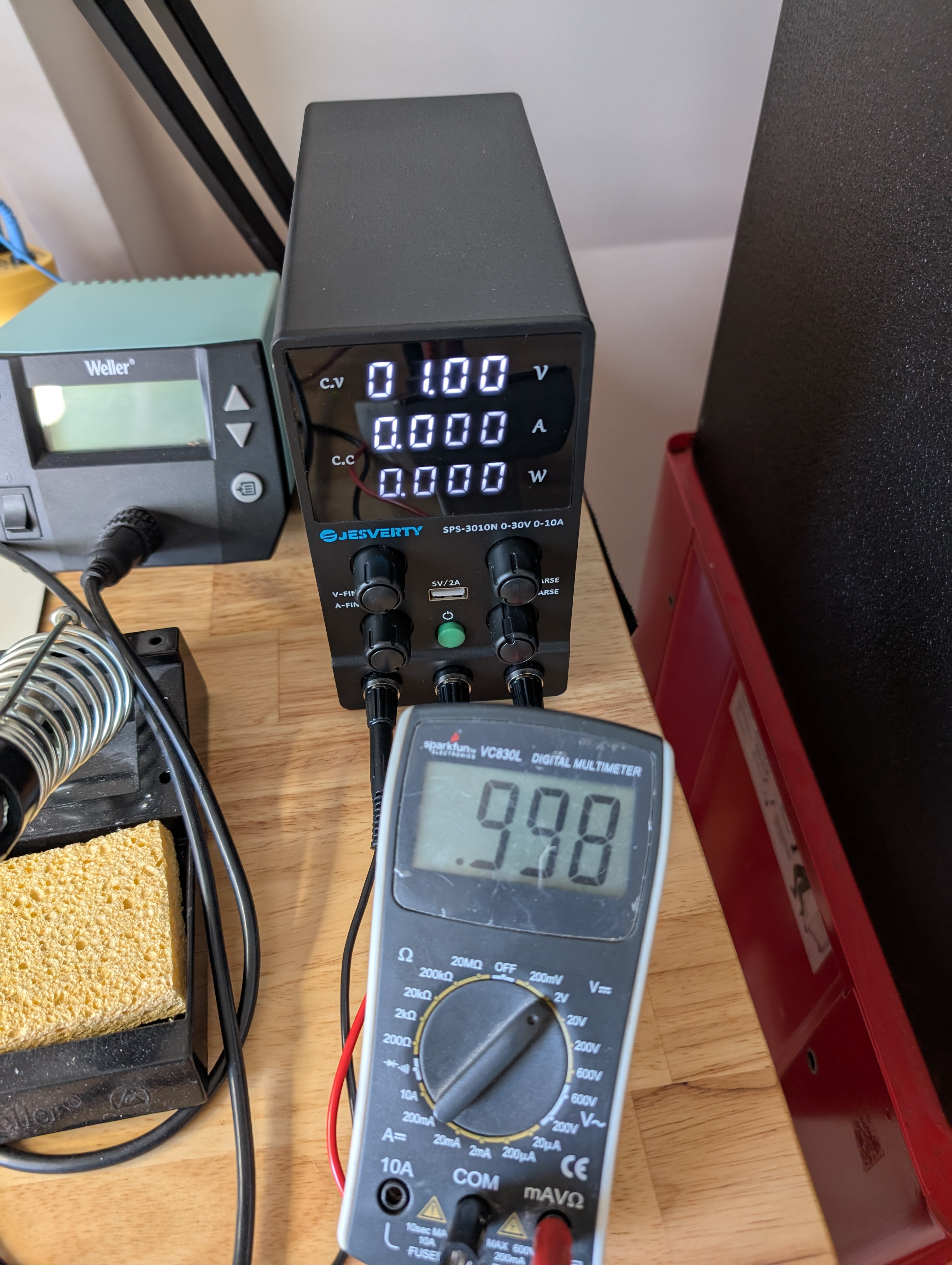 A small black power supply with three numeric displays: 01.00 V, 00.00 A, 00.00 W. In front of the supply is a multimeter reading .998.