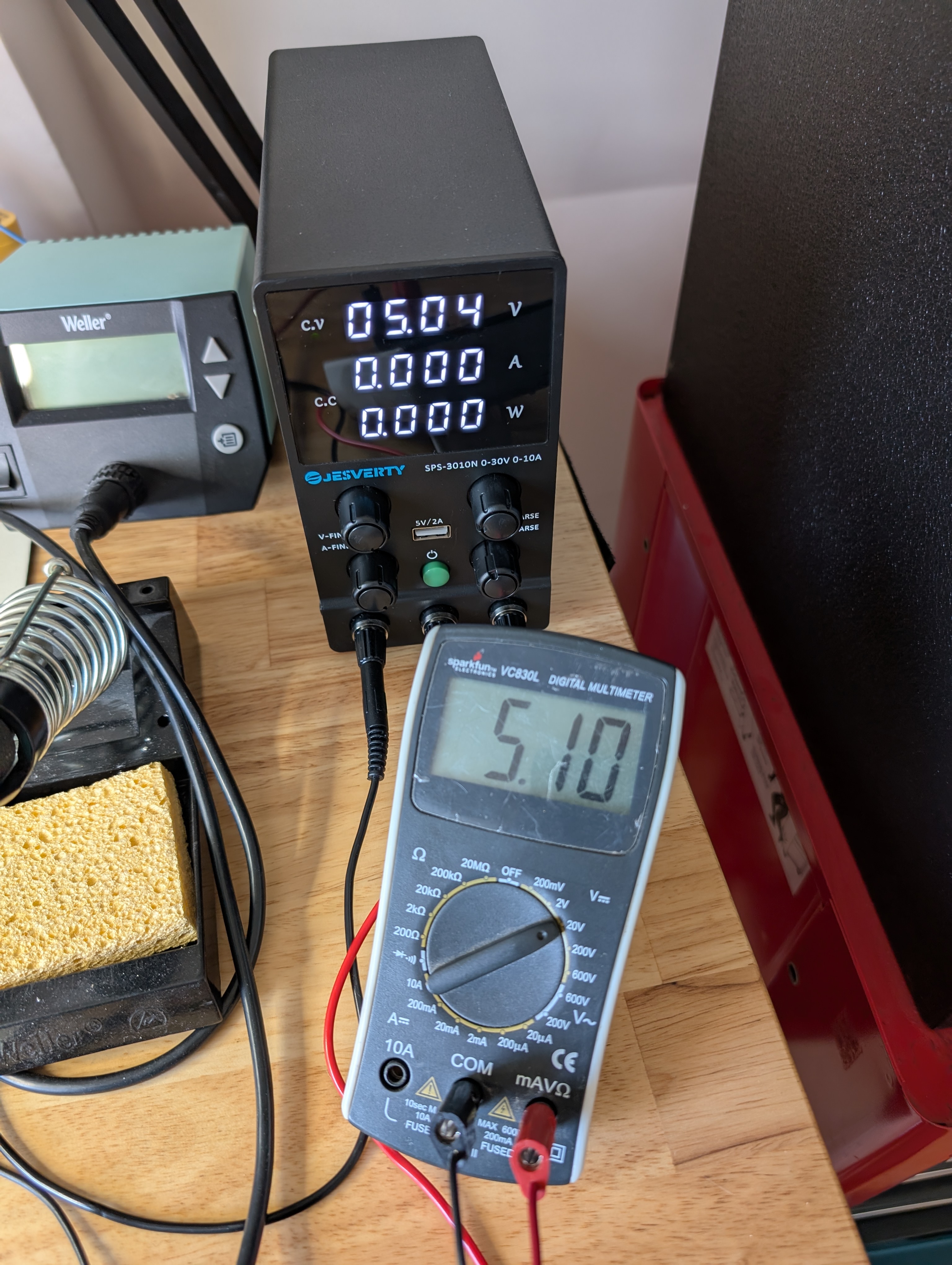 A small black power supply with three numeric displays: 05.04 V, 00.00 A, 00.00 W. In front of the supply is a multimeter reading 5.10.