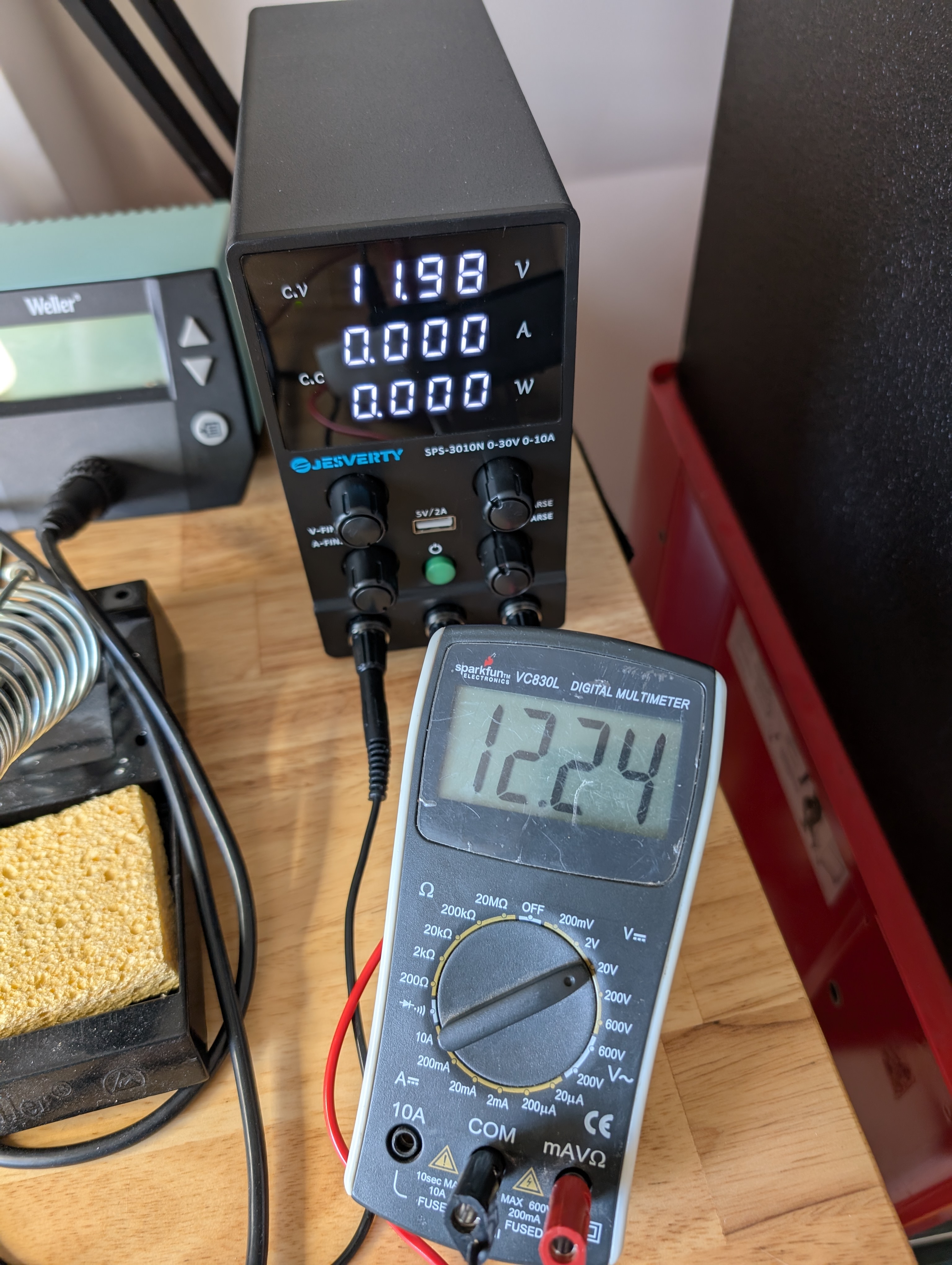 A small black power supply with three numeric displays: 11.98 V, 00.00 A, 00.00 W. In front of the supply is a multimeter reading 12.24.