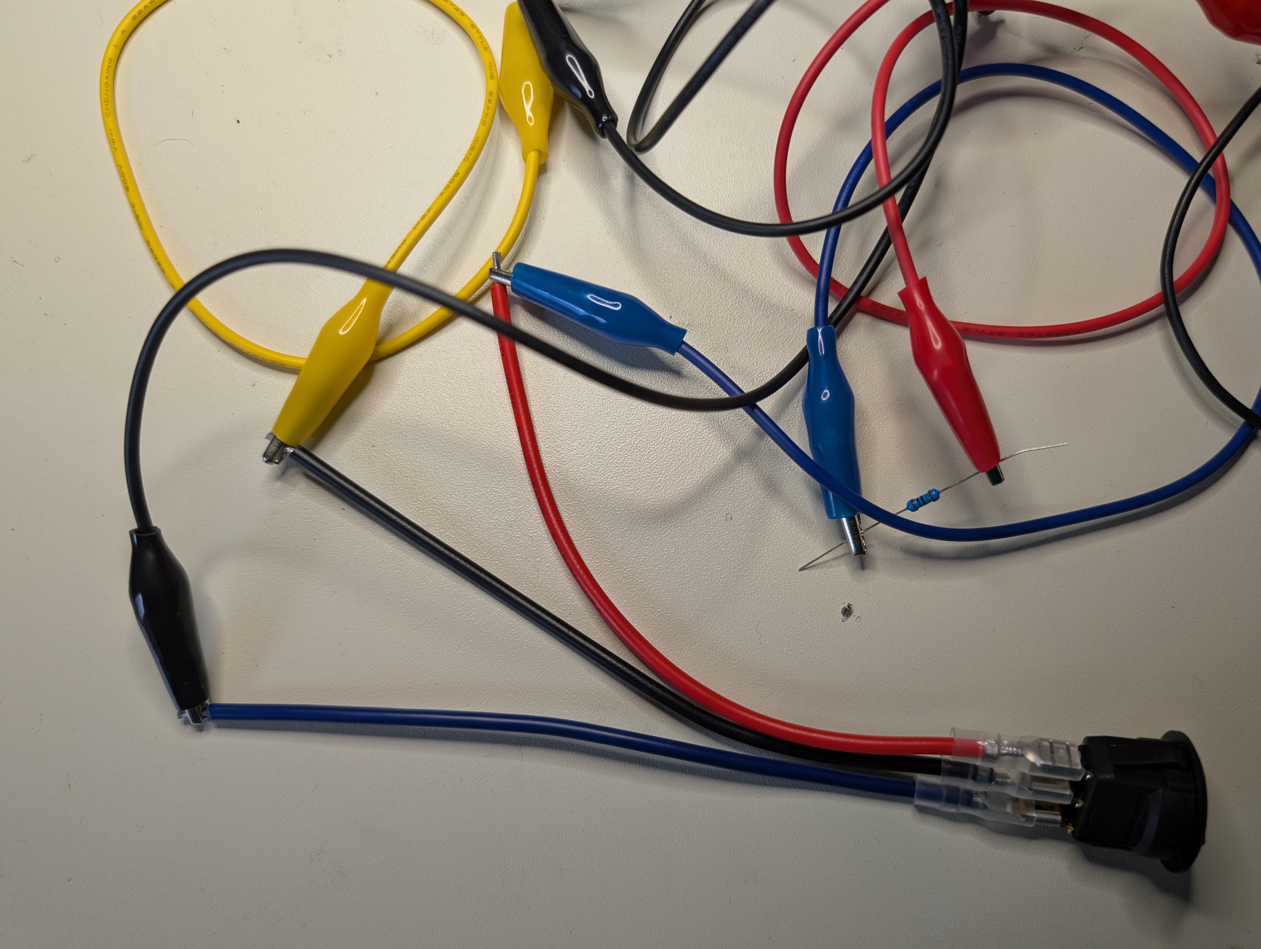 A black plastic switch with three leads, blue, black, and read. There are blue, yellow, red, and black small jumper wires in a jumble, with a resistor clipped between a red and blue jumper clip.