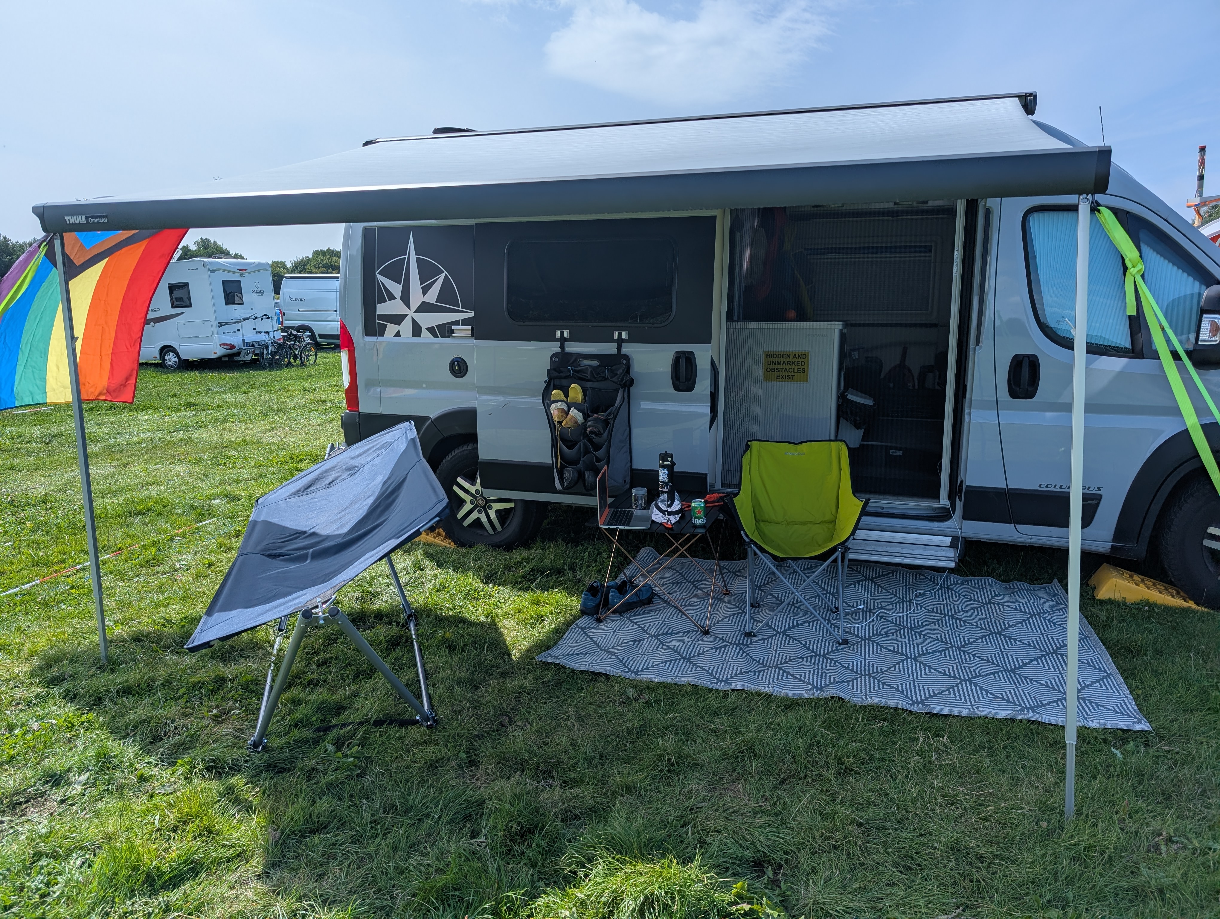 a picture of a grey camper van, our jean-luc, with the awning open. there is a green camp chair, a small table, and a hammock set up