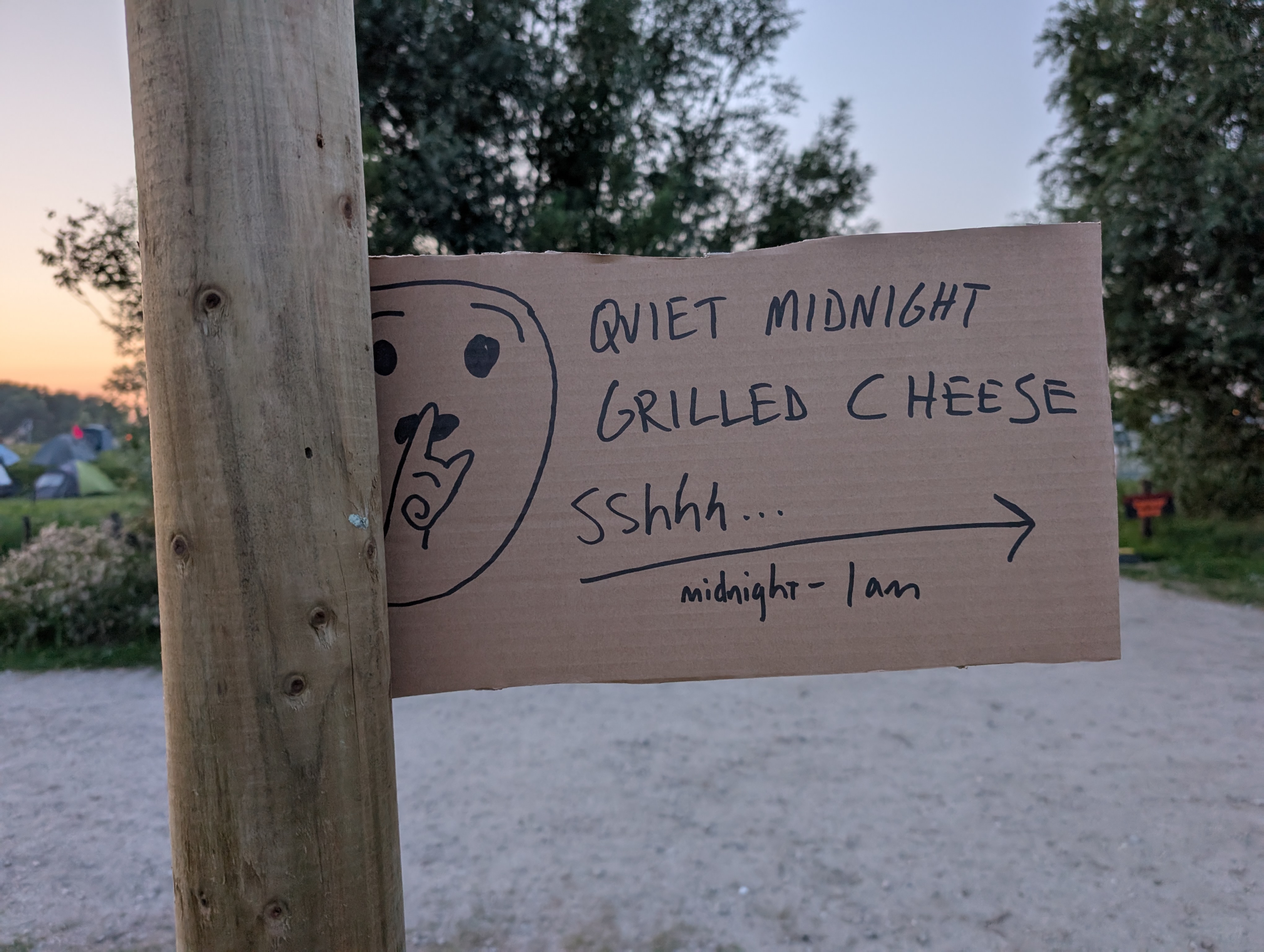 a hand-lettered cardboard sign with a hand-drawn "quiet emoji," partially obscured by the wood post the sign is attached to. the text says "Shhh.... Midnight Tosti, with an arrow to the right.