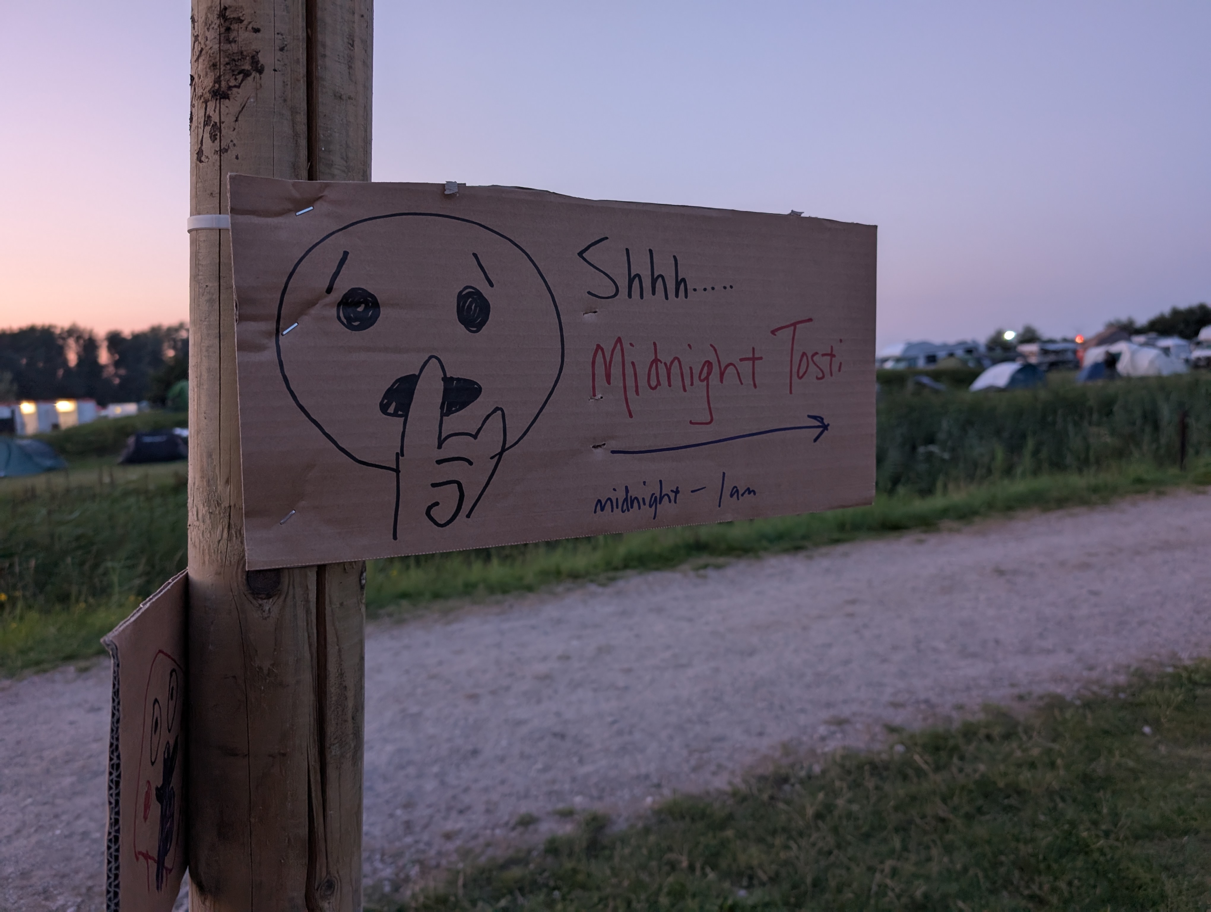 a hand-lettered carboard sign is attached to a wood post. there is a hand drawn "quiet" emoji and the text says "Shhhh.... Midnight Tosti" with an arrow to the right