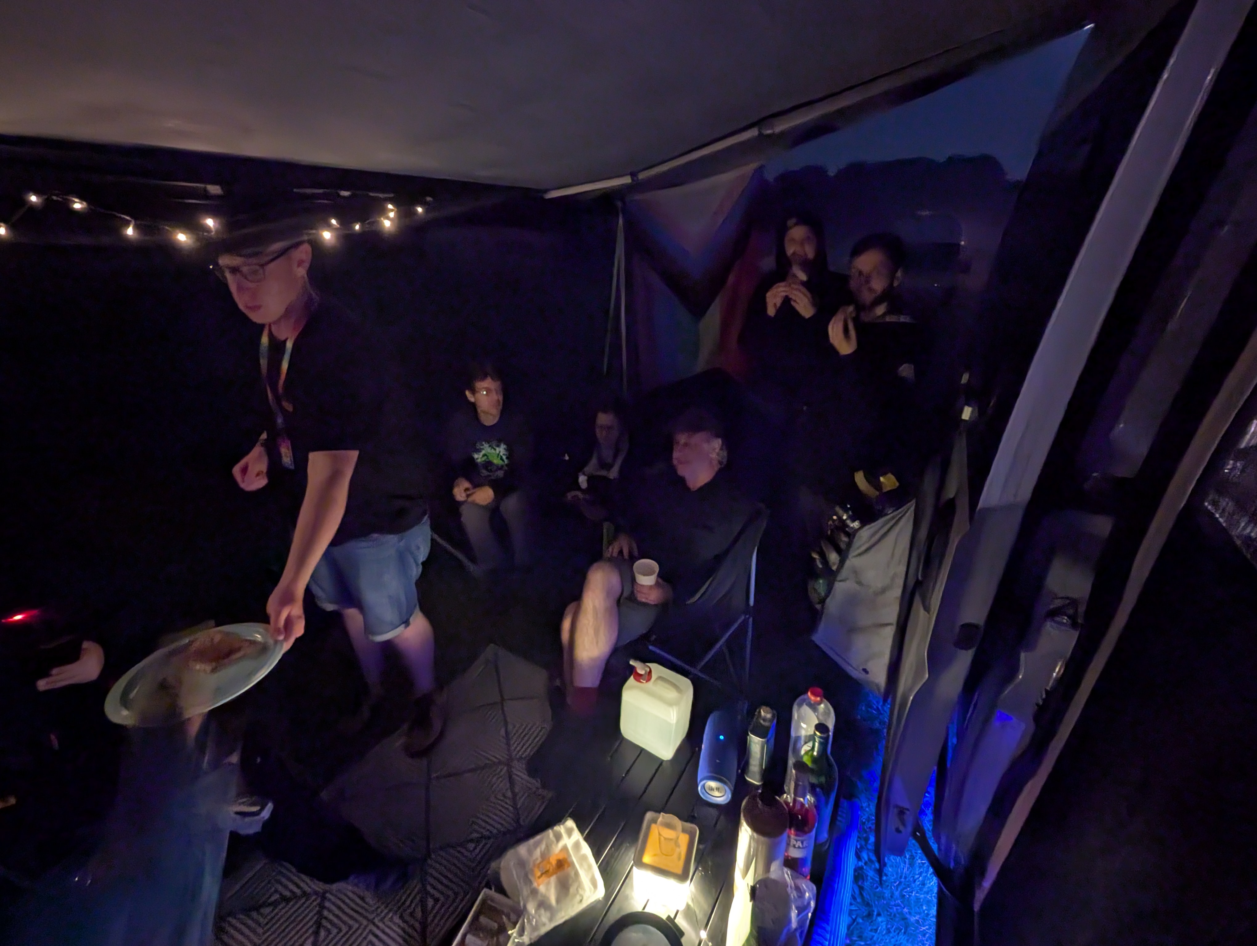 a group of people under a campervan awning at night, eating hot cheese sandwiches. there is a person in the foreground handing a light green plate with a golden tosti on top to someone out of the picture
