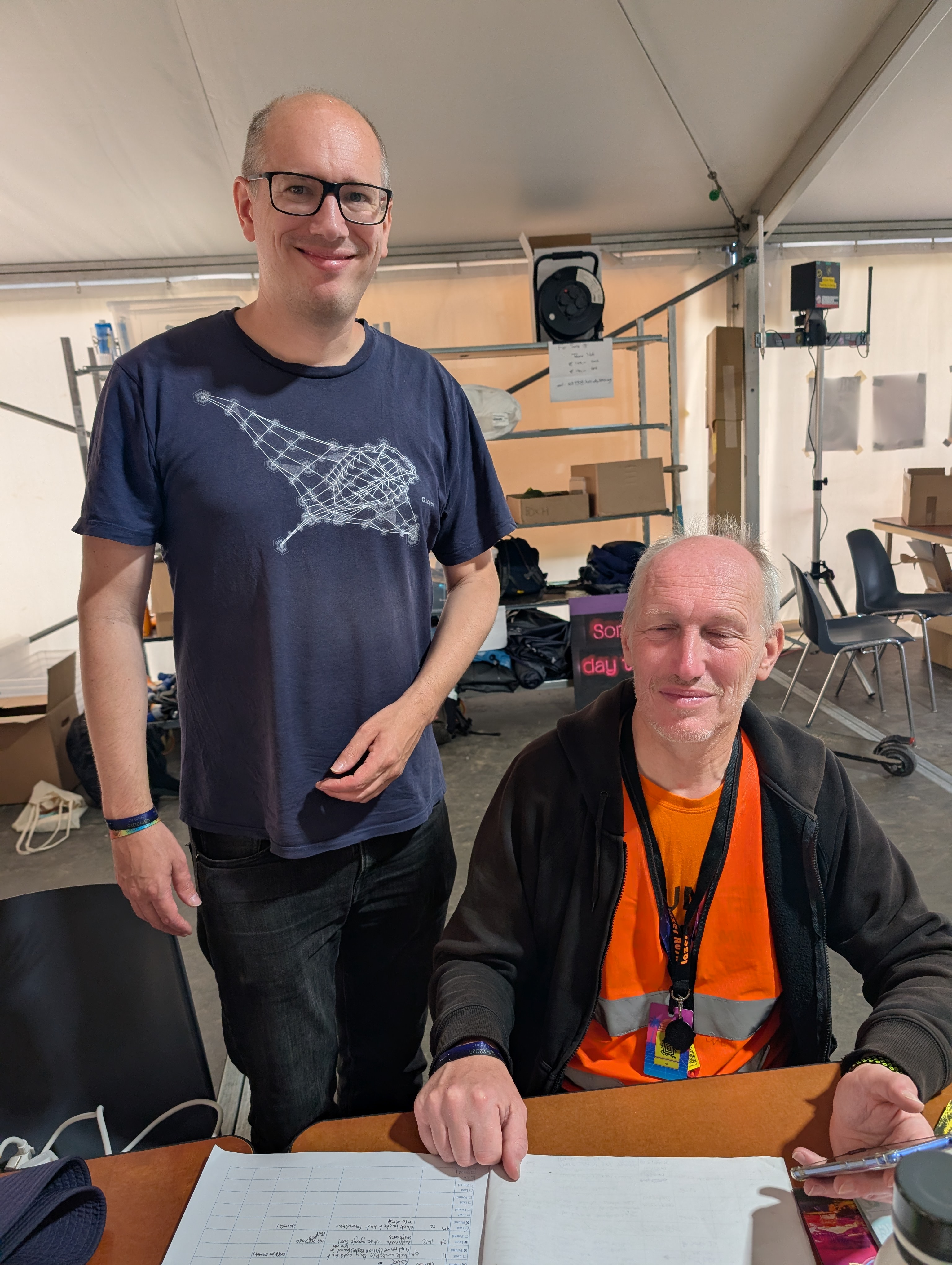two casually dressed men, one standing wearing a blue t-shirt and the other seated and wearing an orange hi-viz jacket
