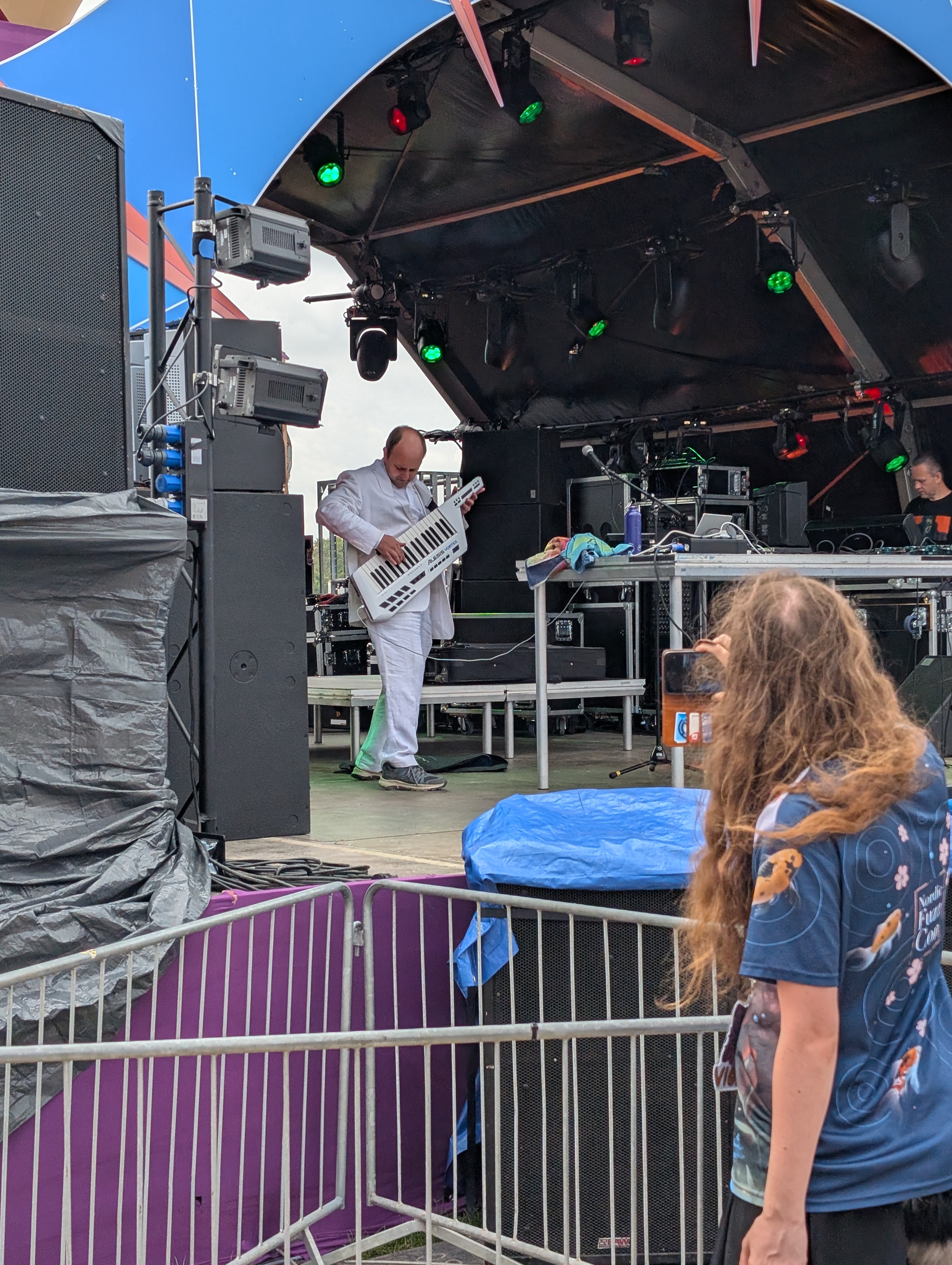 a man in a white suite is playing a white "keytar" on a stage. the back of a person with long unkept hair wearing a blue shirt is in the foreground