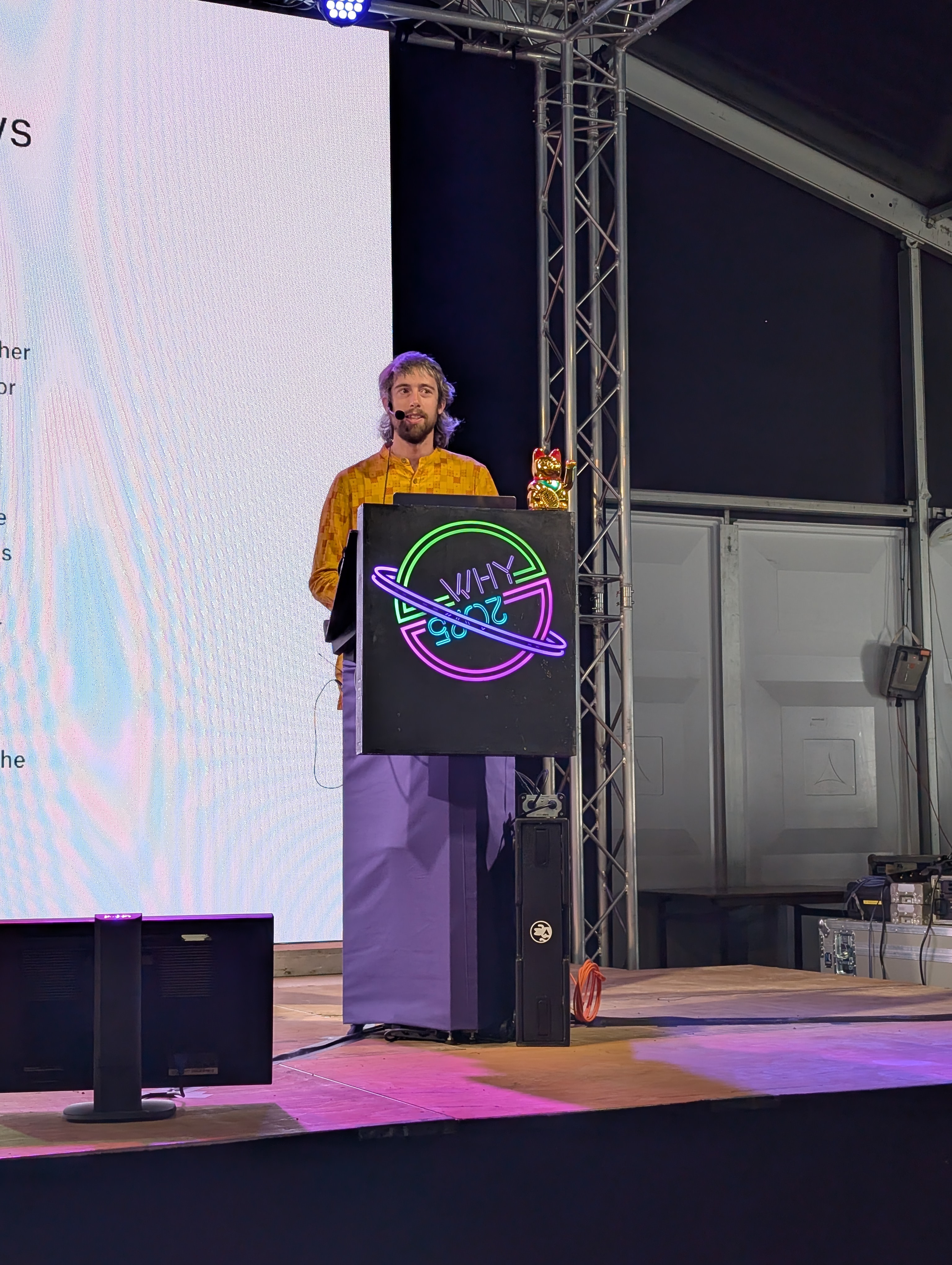 a young-looking bearded person wearing a yellow shirt stands behind a lecturn with the why2025 logo on front
