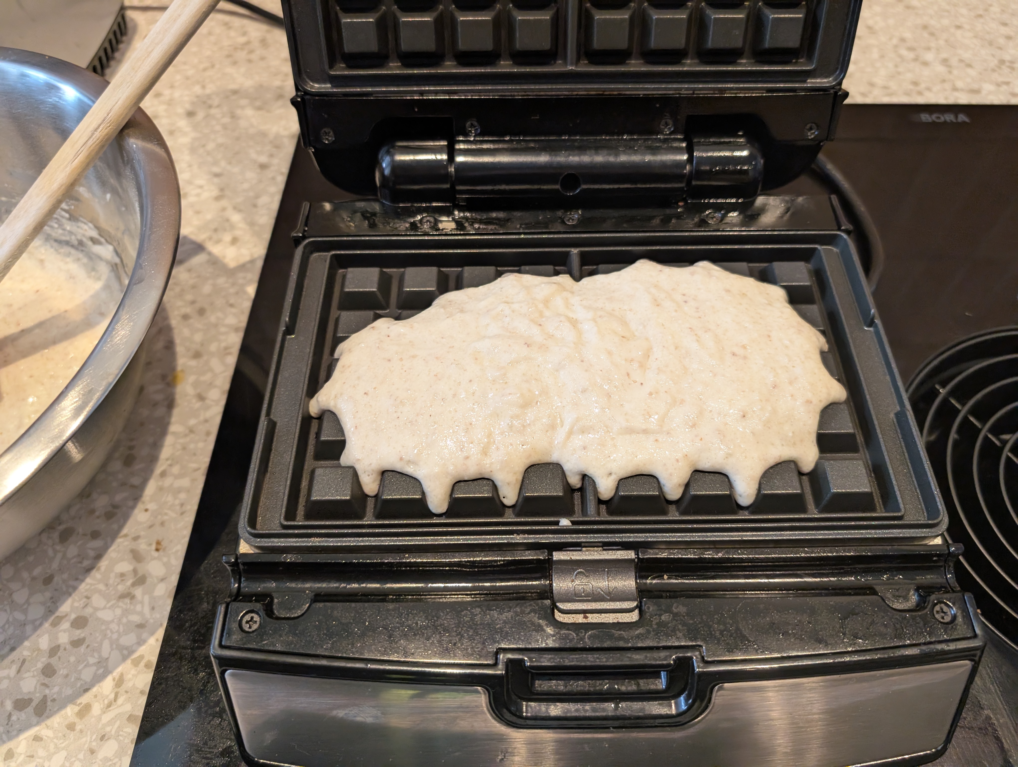 a close up of an oblong patch of off-white batter on a black waffle iron. the edge of a metal mixing bowl containg more batter, with a wooden handle sticking out, is on the left. the waffle iron is on a black glass cooktop and a vent is visible to the right. 
