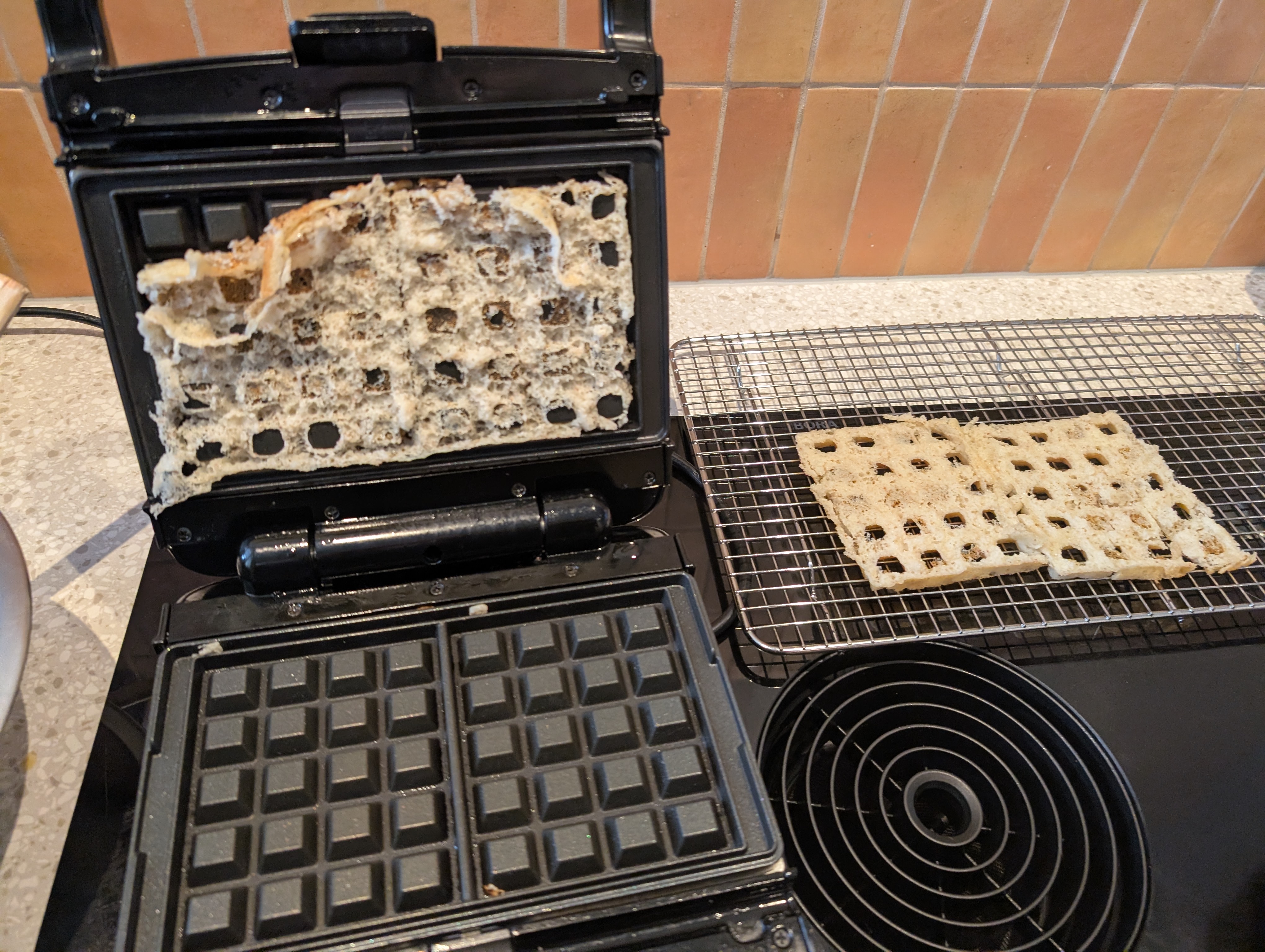 the same waffle iron as in the first picture, but farther out. the bottom grid is clear (some crumbs are visible), but the top grid has half a torn waffle stuck to it. there is the bottom half of the waffle on a cooling rack to the right.