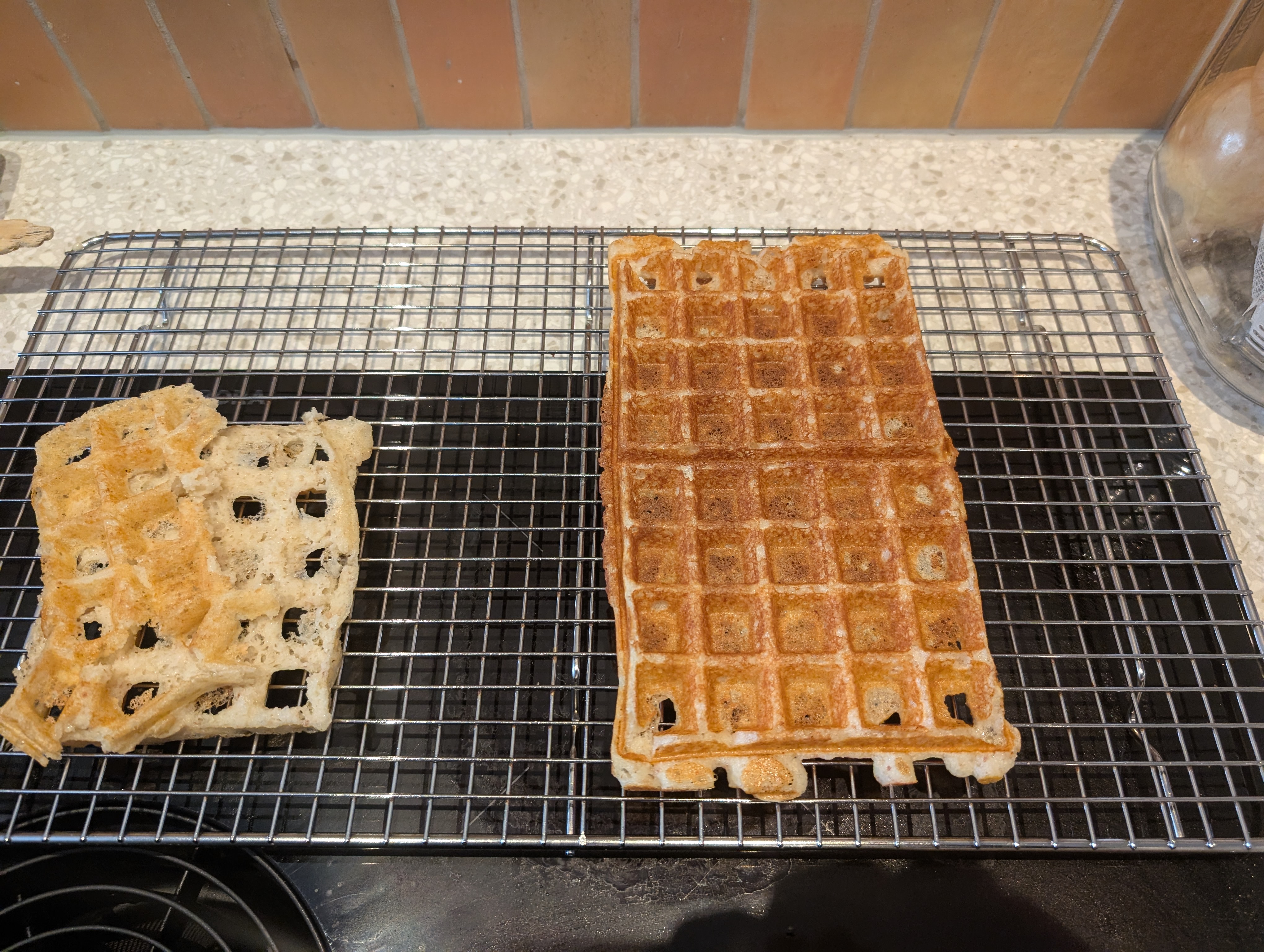 the same waffle from the previous picture on a silver wire cooling rack with a cross-ways grid of about 1 cm pitch. the waffle is rotated 90 degrees so the long side is extending from the foreground to the background. parts of the first torn waffle are on the left of the cooling rack. 