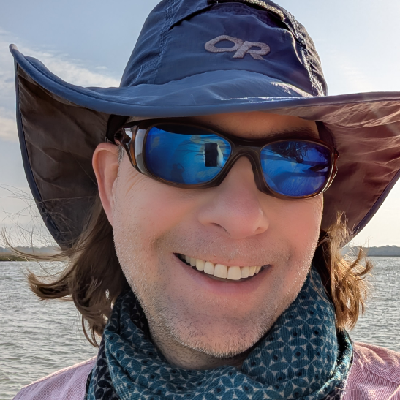 me wearing blue tinted bolle sunglasses with brown frames, a blue wide brimmed sun hat, and a blue scarf over a pink shirt